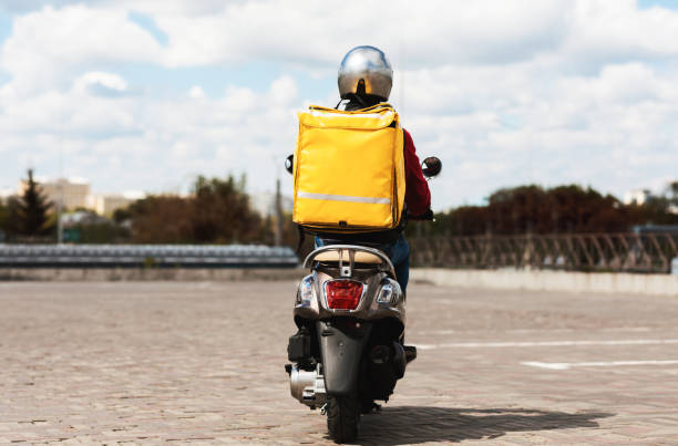 Delivery Service. Courier With Yellow Backpack Riding Scooter Back To Camera In Urban Area Delivering Food In City. Copy Space